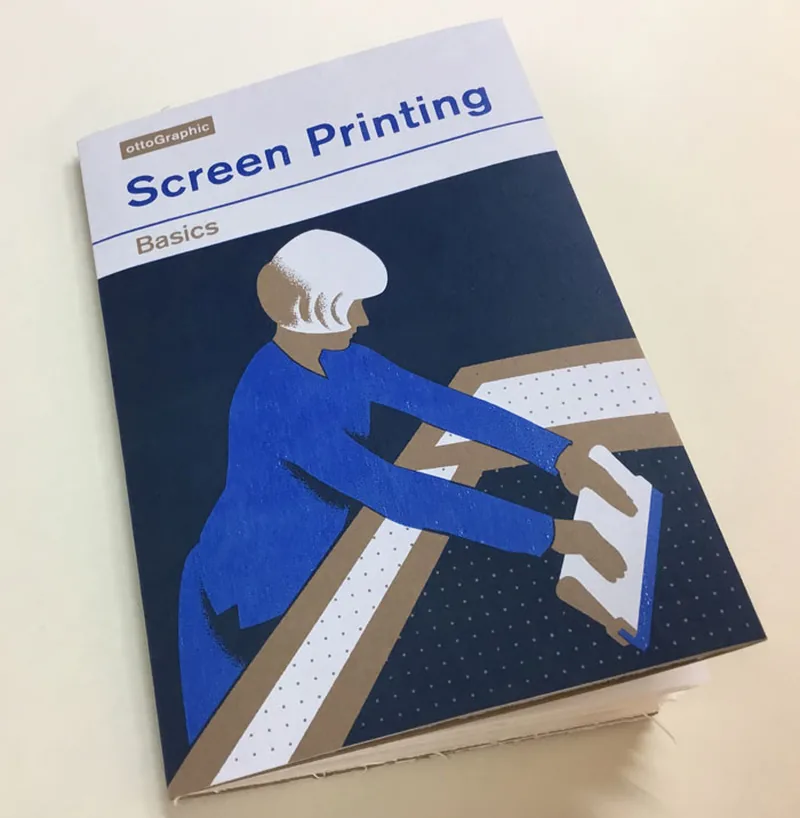 The printed booklet Screen Printing Basics lying closed on a table, its cover showing a figure pulling a squeegee across a tilted screen.
